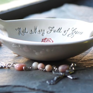May include: White ceramic dish with the words "Hail Mary Full of Grace" and a red rose design. A pink and white rosary with a silver cross is resting on a gray surface.