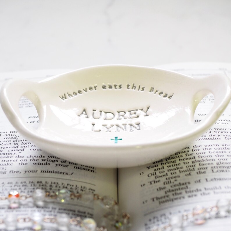 May include: White ceramic dish with two handles, featuring the inscription "Whoever eats this Bread" and the names "Audrey Lynn" in a decorative font. A small turquoise cross is centered below the names.