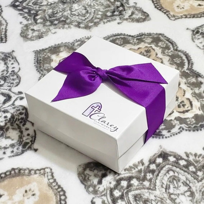 May include: A white gift box with a purple ribbon and bow. The box has a logo that says "Clarcey Clayworks" with a cross symbol.