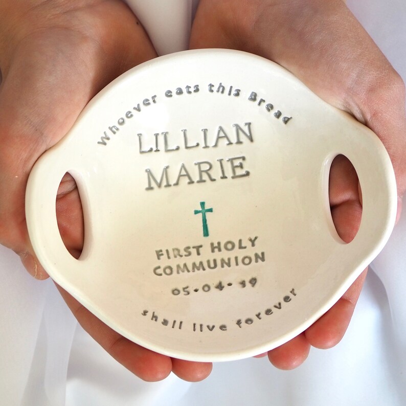 May include: White ceramic dish with two handles, featuring the text "Whoever eats this bread Lillian Marie First Holy Communion 05.04.19 shall live forever" in silver lettering. A teal cross is centered above the text.