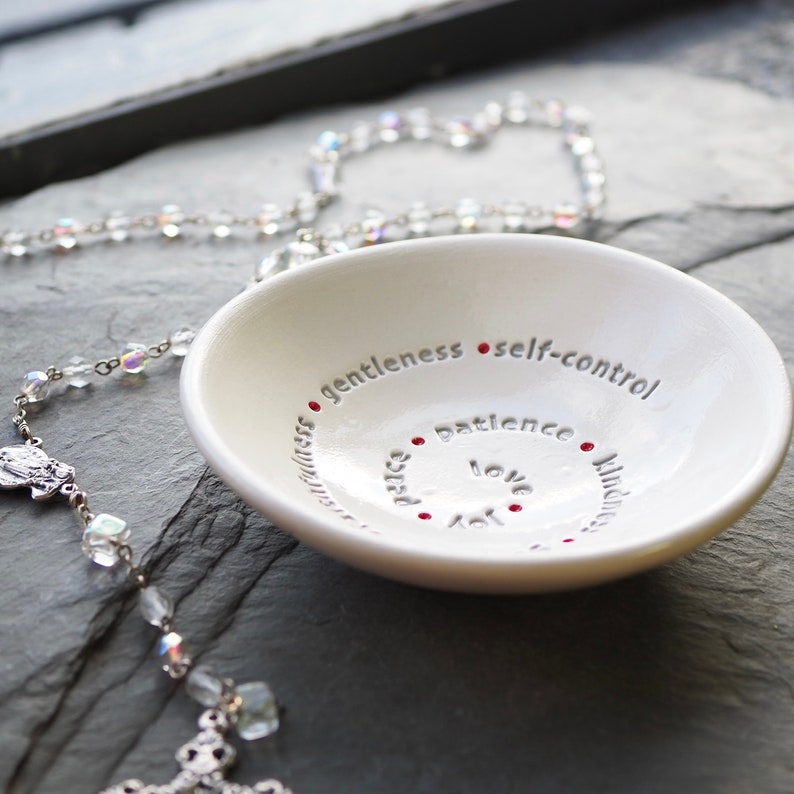 Peut inclure: Un plat en c&eacute;ramique blanche avec les mots "joie, paix, amour, patience, gentillesse, douceur, ma&icirc;trise de soi" inscrits sur le bord. Les mots sont dispos&eacute;s en cercle et s&eacute;par&eacute;s par des points rouges. Un chapelet argent&eacute; est pos&eacute; sur une surface gris fonc&eacute; &agrave; c&ocirc;t&eacute; du plat.