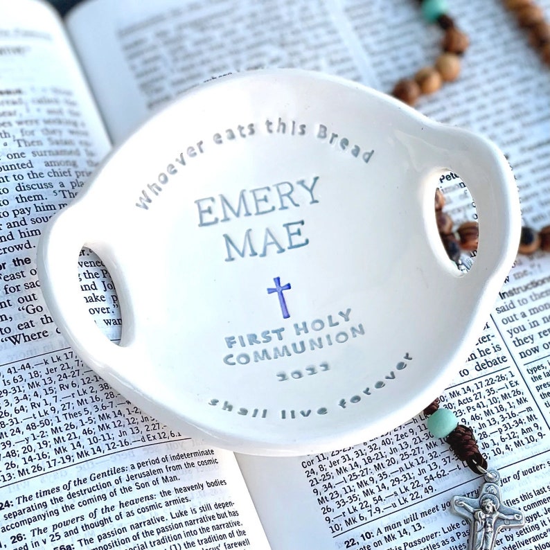 May include: A white ceramic dish with two handles, featuring the text "Whoever eats this Bread Emery Mae First Holy Communion 2023 shall live forever". The dish has a blue cross and the text is in a light blue color.