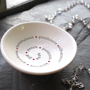 Peut inclure: Plat en c&eacute;ramique blanche avec un motif en spirale de mots comprenant "amour", "joie", "paix", "patience", "gentillesse", "fid&eacute;lit&eacute;", "douceur" et "ma&icirc;trise de soi". Les mots sont en gris avec des points rouges entre chaque mot.