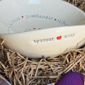 May include: A white ceramic bowl with the words "gentleness, selflessness, kindness" and "sponsor 2022" inscribed on the inside. A red heart symbol is next to the year.