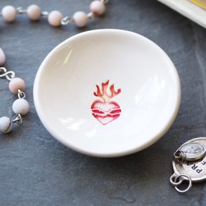 May include: White ceramic dish with a red and yellow heart with flames design. The dish is perfect for holding jewelry or other small items.