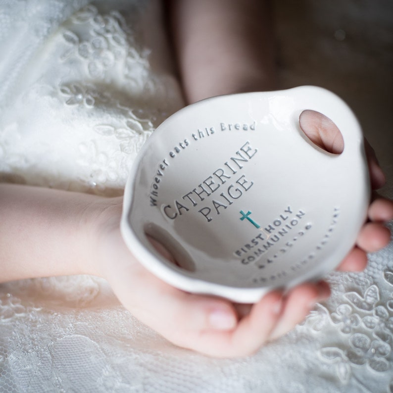 May include: White ceramic dish with handles, inscribed with the text "Whoever eats this Bread... Catherine Paige" and a cross. The text "First Holy Communion" is also inscribed on the dish.