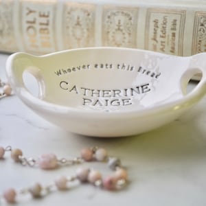 May include: White ceramic dish with two handles. The dish has the text "Whoever eats this bread Catherine Paige" inscribed on the bottom.