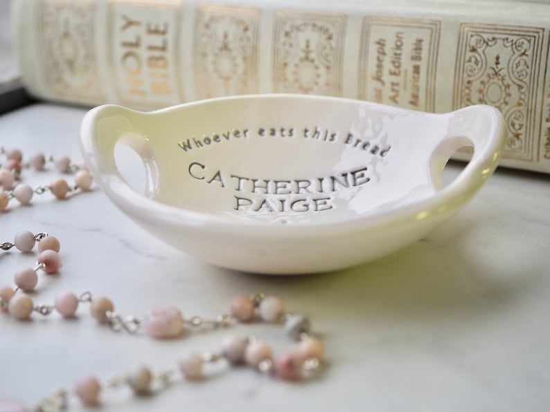 May include: White ceramic dish with two handles. The dish has the text "Whoever eats this bread Catherine Paige" inscribed on the bottom.