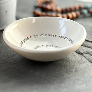 A white ceramic bowl featuring the nine Fruits of the Holy Spirit. The side view shows the depth of the bowl and a wooden rosary sits in the background.