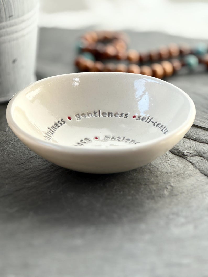 A white ceramic bowl featuring the nine Fruits of the Holy Spirit. The side view shows the depth of the bowl and a wooden rosary sits in the background.