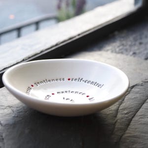 A white ceramic bowl featuring the nine Fruits of the Holy Spirit is on a slate table top with a window in the back