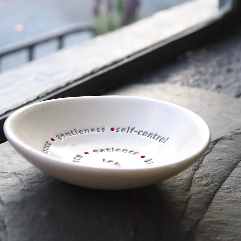 A white ceramic bowl featuring the nine Fruits of the Holy Spirit is on a slate table top with a window in the back