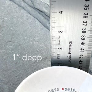 Part of a white ceramic bowl featuring the nine Fruits of the Holy Spirit is shown next to a ruler that reads a depth of 1 inch.