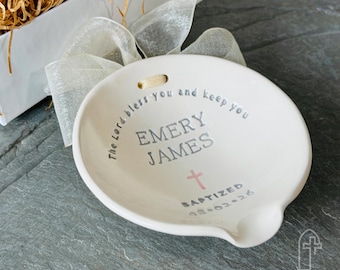 Op de afbeelding: Een witte keramische lepelhouder met een zilveren lint en de tekst "The Lord bless you and keep you", "EMERY JAMES", "BAPTIZED" en "08.02.26" in zilveren letters, met een klein roze kruisje onder de naam.