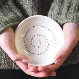 A woman is holding a white ceramic bowl featuring the nine Fruits of the Holy Spirit