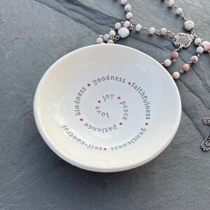 A white ceramic bowl featuring the nine Fruits of the Holy Spirit sits on a slate top with a pink rosary near it.