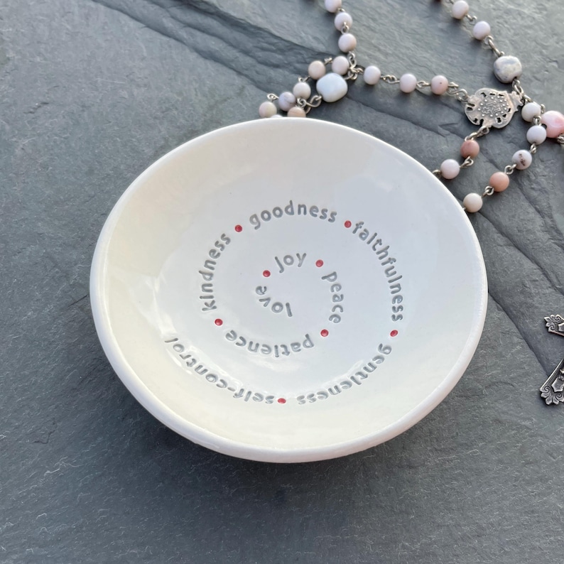 A white ceramic bowl featuring the nine Fruits of the Holy Spirit sits on a slate top with a pink rosary near it.