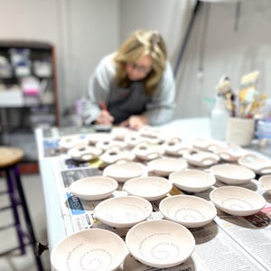 A blonde haired women stands at a table with several handmade bowls on it.