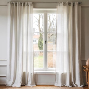 May include: White sheer curtains hang in front of a window with a view of trees and a house in the distance. The curtains are gathered at the top and have a simple, elegant design.