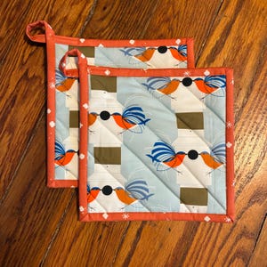 May include: Two square quilted pot holders with a light blue background featuring a repeating pattern of birds wearing sunglasses. The pot holders have an orange border with white polka dots and a loop for hanging.