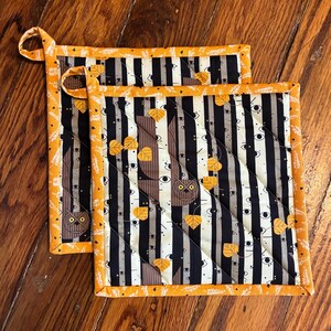 May include: Two square quilted pot holders with a Halloween theme. The design features vertical black and white stripes with owl and eye patterns, accented with orange and yellow leaves. The edges are trimmed with orange fabric.