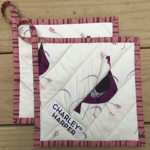 May include: Two square quilted pot holders with a bird design. The pot holders have a white background with a diagonal stitch pattern and a bird illustration in shades of purple. The edges are trimmed with a dark pink fabric. The label reads "CHARLEY HARPER".