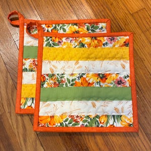 May include: Two square quilted pot holders featuring horizontal stripes of floral fabric in orange, yellow, and green, interspersed with white fabric strips patterned with wheat stalks. Each piece is finished with a solid orange fabric binding.
