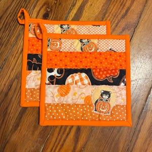 May include: Two square quilted pot holders featuring a patchwork design of orange, black, and cream fabric strips. The patterns include polka dots, pumpkins, and a black cat sitting on a pumpkin. Each pot holder has a bright orange fabric border.
