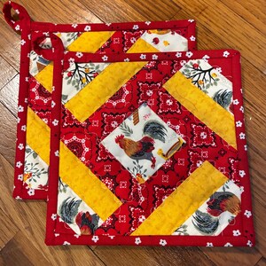 May include: Two square quilted pot holders featuring a patchwork design of red bandana print fabric, yellow textured fabric, and white fabric with a rooster and floral pattern. Each pot holder has a red border with a small white flower print.