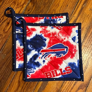 May include: Two square quilted pot holders featuring a red, white, and blue tie-dye pattern with the Buffalo Bills logo and the text BILLS. Each pot holder has a dark blue border with white polka dots and a small fabric loop for hanging.