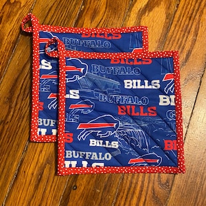 May include: Two blue quilted pot holders with red trim and hanging loops. The pot holders feature the words "BUFFALO BILLS" in white and red, along with a buffalo logo and a stadium graphic.