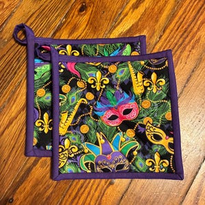 May include: Two square quilted pot holders featuring a black background with a vibrant Mardi Gras pattern. The design includes colorful masquerade masks, gold fleur-de-lis symbols, yellow saxophones, green peacock feathers, and gold beads.