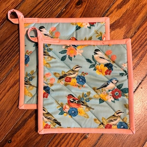 May include: Two square quilted pot holders with a light blue background. The pot holders feature a floral and bird pattern in various colors, including red, yellow, and blue. The edges are trimmed with a peach-colored fabric and have a loop for hanging.