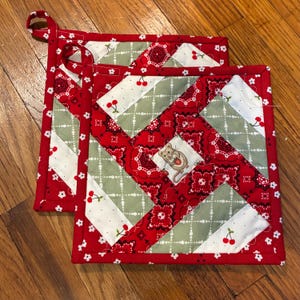 May include: Two square quilted pot holders featuring a patchwork design of red bandana print, sage green fabric with white dots, and white fabric with red cherry patterns. A small illustration of a mouse holding a red cherry is centered.