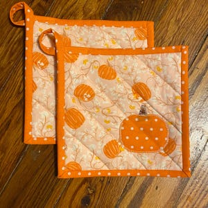 May include: Two quilted orange and white pot holders. The pot holders have a pumpkin pattern with orange trim and white polka dots. One pot holder has a loop for hanging.