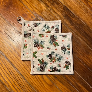 May include: Two beige pot holders with a pine cone and holly berry pattern. The pot holders have a loop for hanging.