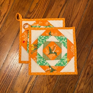 May include: Two orange and white quilted pot holders with a floral pattern. The pot holders have a white background with orange and green squares and a white polka dot border.
