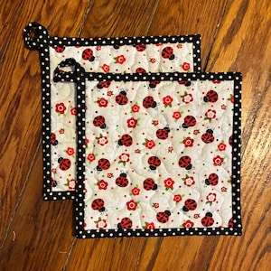 May include: Two square quilted potholders with a white background featuring red ladybugs and flowers. The potholders have a black polka dot border and a loop for hanging.