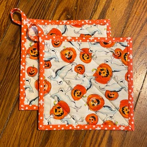 May include: Two square Halloween-themed pot holders. The white fabric is patterned with orange jack-o'-lanterns and gray ghosts. The edges are trimmed with orange polka dot fabric. One pot holder has a fabric loop for hanging.