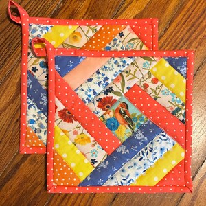 May include: Two square quilted pot holders with a patchwork design. The patchwork includes floral and polka dot patterns in yellow, orange, blue, and white. A red polka dot border and hanging loop complete the design. The pot holders are on a wooden surface.