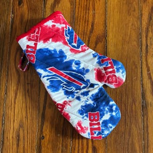 May include: A tie-dye oven mitt in red, white, and blue, featuring the Buffalo Bills logo and the word "Bills" in bold letters. The mitt has a red trim and a loop for hanging.