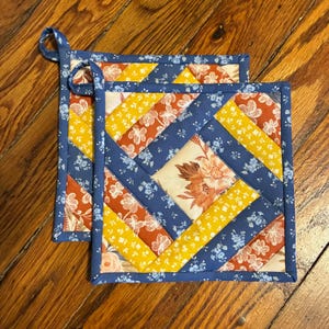 May include: Two handmade quilted pot holders. Each square pot holder features a patchwork design with floral and geometric patterns in shades of blue, yellow, orange, and cream. A blue loop is attached for hanging.