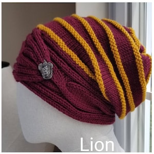 May include: A maroon and gold knitted hat with a lion crest. The hat has a cable knit design and a striped pattern. The hat is on a mannequin head.