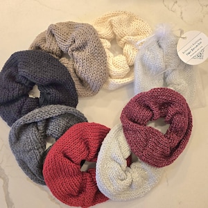 May include: A collection of handmade knitted hair scrunchies in various colors, including gray, navy, red, cream, and burgundy. One scrunchie is packaged in a clear bag with a tag that reads "Rebecca's Knitting Hair Scrunchie."