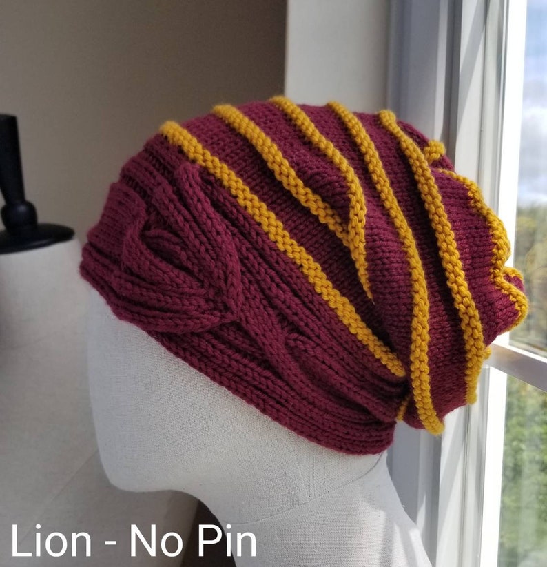 May include: A maroon knitted beanie with a cable knit pattern and yellow stripes. The beanie is displayed on a white mannequin head. "Lion - No Pin" is written below the beanie.