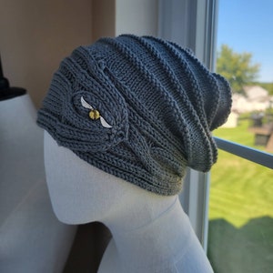 May include: A gray knitted beanie with a cable knit pattern and a gold and white embroidered design of a snitch.