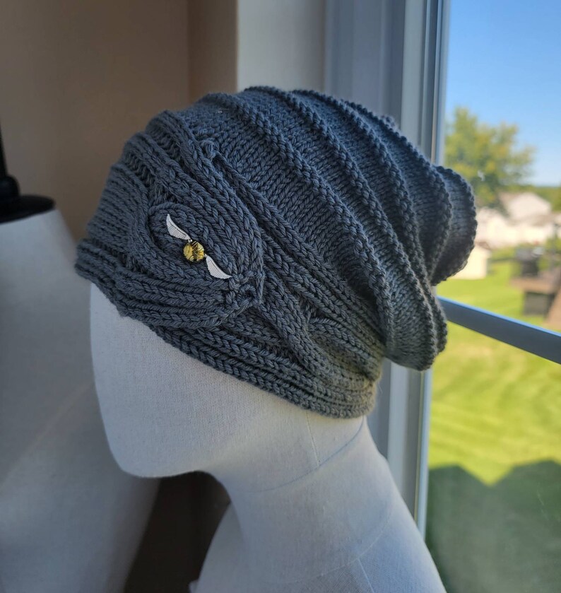 May include: A gray knitted beanie with a cable knit pattern and a gold and white embroidered design of a snitch.