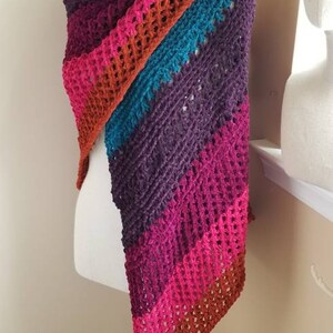May include: A colorful, handmade crochet shawl draped over a mannequin. The shawl features a gradient of colors, including teal, purple, pink, orange, and red. The open weave design adds texture and visual interest to this cozy accessory.