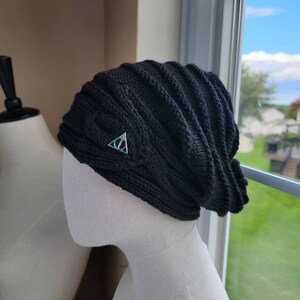 May include: Black knitted beanie with a silver triangle symbol on the front. The symbol has a circle inside the triangle.