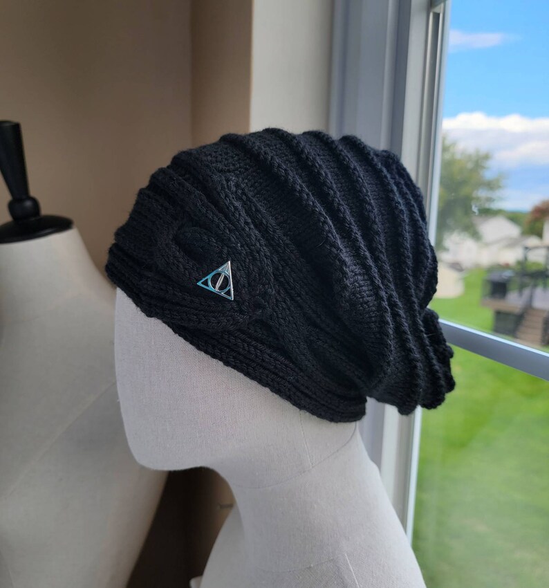 May include: Black knitted beanie with a silver triangle symbol on the front. The symbol has a circle inside the triangle.
